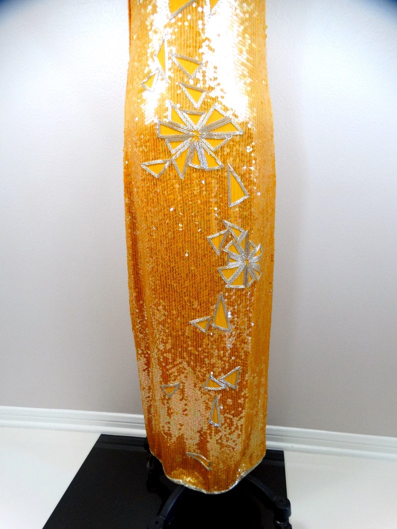 K&ouml;nnte beinhalten: Ein langes, goldenes Paillettenkleid mit einem silbernen geometrischen Muster. Das Kleid hat ein figurbetontes Oberteil und einen flie&szlig;enden Rock.