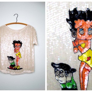 Peut inclure: Un chemisier blanc à paillettes avec une image de dessin animé de Betty Boop et un personnage du dessin animé. Le chemisier a des manches courtes et un col rond.