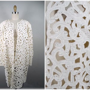 Puede incluir: Una chaqueta blanca de ganchillo con un delicado patrón floral y detalles de cuentas. La chaqueta está abierta en la parte delantera y tiene mangas largas.