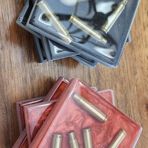 May include: Set of four square coasters with a red and black resin base. Each coaster features a clear epoxy top with gold-colored bullet casings embedded inside.
