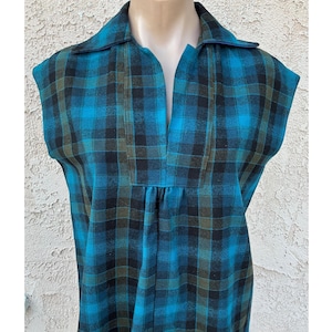 May include: A sleeveless, vintage-style dress in a teal, black, and olive green plaid pattern. The dress features a collared neckline and a V-neck opening. The fabric appears to be a soft, woven material.