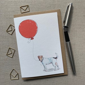 Può includere: Un biglietto bianco con un palloncino rosso e un'illustrazione in bianco e nero di un cane che tiene il filo del palloncino. Il biglietto è su una superficie marrone.