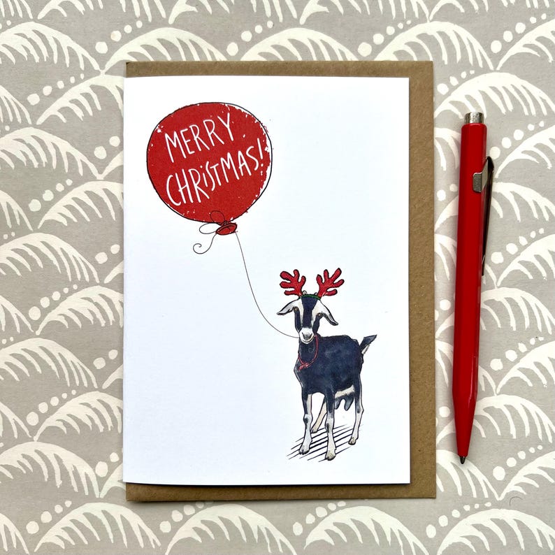 Puede incluir: Tarjeta de Navidad con una cabra negra con cuernos de reno rojos y un collar rojo, sosteniendo un globo rojo con "&iexcl;FELIZ NAVIDAD!". Un bol&iacute;grafo rojo est&aacute; a la derecha. La tarjeta est&aacute; sobre un fondo estampado.