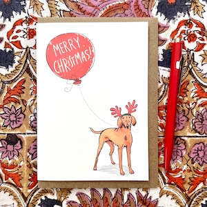 May include: A Christmas card featuring a dog wearing reindeer antlers and holding a balloon with the words "MERRY CHRISTMAS!" in a red bubble. The card is in a brown envelope, with a red pen on the side.