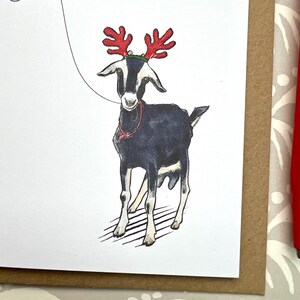 Puede incluir: Una tarjeta de Navidad con una cabra en blanco y negro con cuernos de reno rojos y un collar rojo. La cabra est&aacute; sobre un fondo blanco con un borde marr&oacute;n.