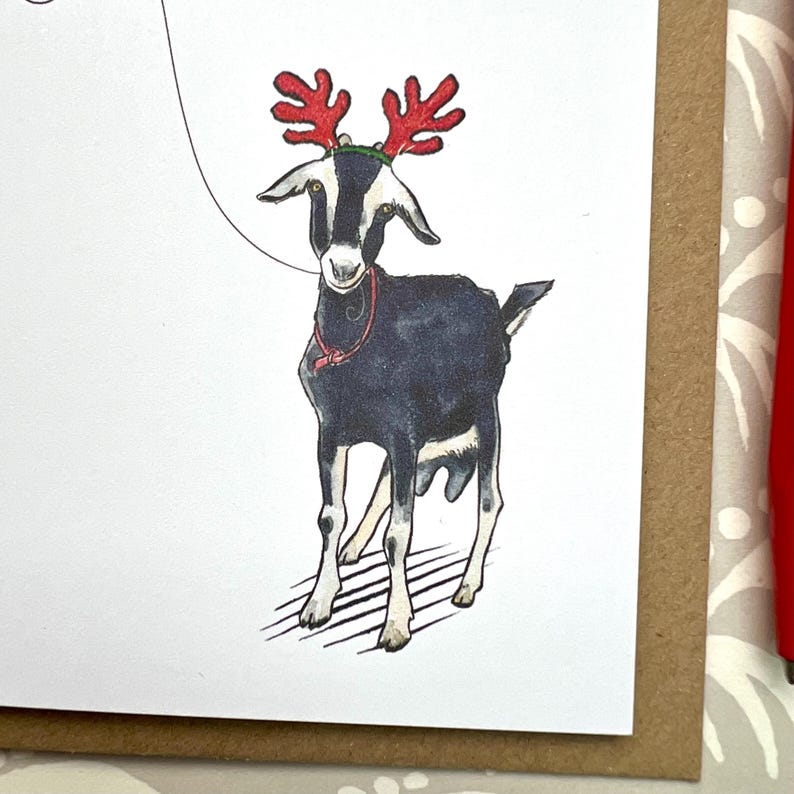 Puede incluir: Una tarjeta de Navidad con una cabra en blanco y negro con cuernos de reno rojos y un collar rojo. La cabra est&aacute; sobre un fondo blanco con un borde marr&oacute;n.