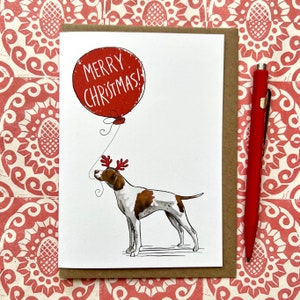Può includere: Biglietto di Natale con un cane con corna di renna e un palloncino rosso con la scritta "MERRY CHRISTMAS!". Il biglietto è su uno sfondo marrone. Una penna rossa è a destra.