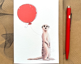 May include: A greeting card featuring a watercolor illustration of a meerkat holding a red balloon. The meerkat is standing on its hind legs and looking directly at the viewer. The card is blank inside for writing a message.