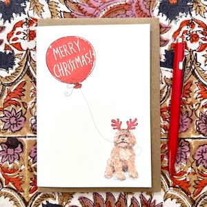 May include: A Christmas card featuring a brown dog wearing red antlers, holding a red balloon with the words "MERRY CHRISTMAS!" in white. The card is on a brown background with a red pen to the right.