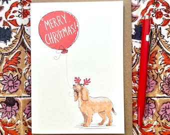 May include: A Christmas card featuring a light brown dog wearing reindeer antlers and looking up at a red balloon. The balloon has the words "MERRY CHRISTMAS!" printed on it. A red pen is next to the card.