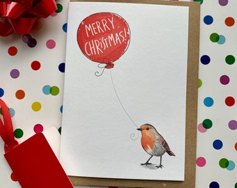 May include: A Christmas card featuring a red balloon with the words "MERRY CHRISTMAS!" and a small bird holding the string. The card is on a kraft paper backing. A red gift tag and bow are in the background.