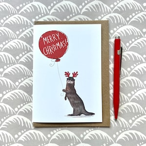 Könnte beinhalten: Weihnachtskarte mit einer Illustration eines Otters mit Rentiergeweih und einem roten Ballon mit der Aufschrift "MERRY CHRISTMAS!". Ein roter Stift liegt neben der Karte. Der Hintergrund ist ein neutrales Muster.