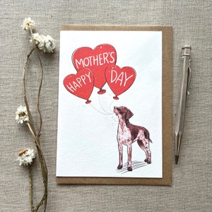 May include: A Mother's Day card featuring a brown and white dog gazing at three red heart-shaped balloons with the words "Happy Mother's Day". The card is on a brown envelope, with a silver pen and dried flowers nearby.