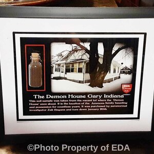 May include: A black framed display featuring a small glass vial with a cork stopper containing soil, a black and white photograph of a house, and text reading The Demon House Gary Indiana with a description of the soil sample's origin.