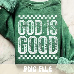 K&ouml;nnte beinhalten: Gr&uuml;nes Sweatshirt mit dem Text "GOD IS GOOD" in verblichener wei&szlig;er Schrift. Das Design beinhaltet ein Schachbrettmuster &uuml;ber und unter dem Text. Die W&ouml;rter sind leicht strukturiert mit Sternakzenten.