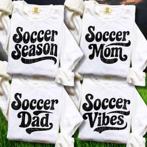 Op de afbeelding: Vier witte shirts met lange mouwen met zwarte tekst. De shirts lezen "Soccer Season", "Soccer Mom", "Soccer Dad" en "Soccer Vibes". De shirts worden gepresenteerd op een groen oppervlak.