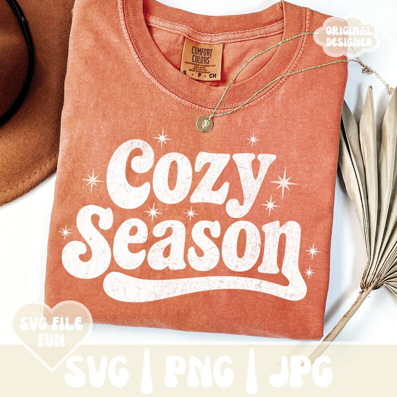 Puede incluir: Una camiseta de color coral con las palabras "Cozy Season" en letras blancas de estilo retro. La camiseta tiene cuello redondo y mangas cortas. Un collar dorado con un colgante est&aacute; drapeado sobre la camiseta. La camiseta est&aacute; doblada y tiene una etiqueta que dice "Comfort Colors".