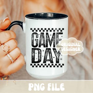 K&ouml;nnte beinhalten: Wei&szlig;er Keramikbecher mit schwarzem Innenraum und Henkel. Der Becher zeigt den Text "GAME DAY" in einer kr&auml;ftigen, verwitterten Schrift, mit einem Schachbrettmuster &uuml;ber und unter dem Text. Der Becher wird von einer Person gehalten.