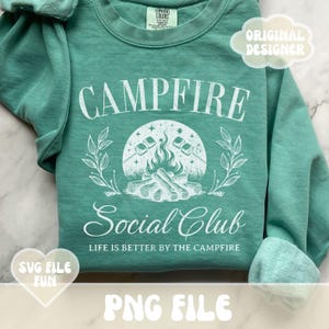Może przedstawiać: Zielony, turkusowy sweter z białym nadrukiem, na kt&oacute;rym widnieje napis "Campfire Social Club" i "Life is better by the campfire". Grafika przedstawia ognisko z pieczeniem pianek nad płomieniami.