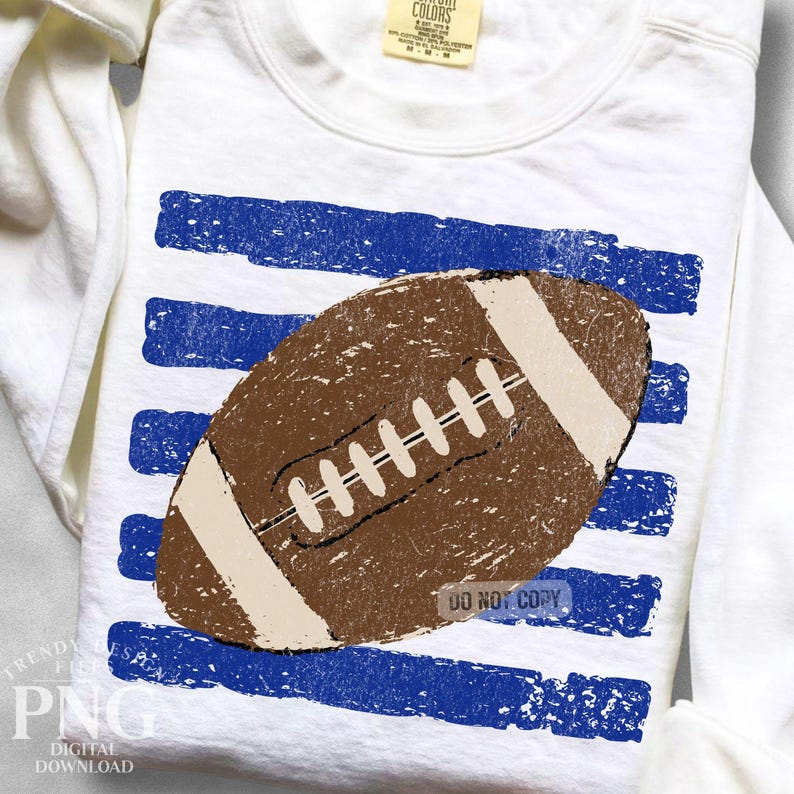 K&ouml;nnte beinhalten: Ein wei&szlig;es Langarm-Sweatshirt mit einer Grafik eines braunen American Footballs mit wei&szlig;en Schn&uuml;rsenkeln und Streifen. Der Football ist &uuml;ber f&uuml;nf horizontalen, abgenutzten blauen Pinselstrich-Streifen zentriert. Das Kleidungsst&uuml;ck hat ein Nackenetikett.