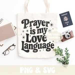Op de afbeelding: Een cr&egrave;mekleurige draagtas met de zwarte tekst "Prayer is my Love Language". De afbeelding bevat een paspoort, een camera, een bril, een notitieboek en groen. Het ontwerp heeft ster-accenten rond de tekst.