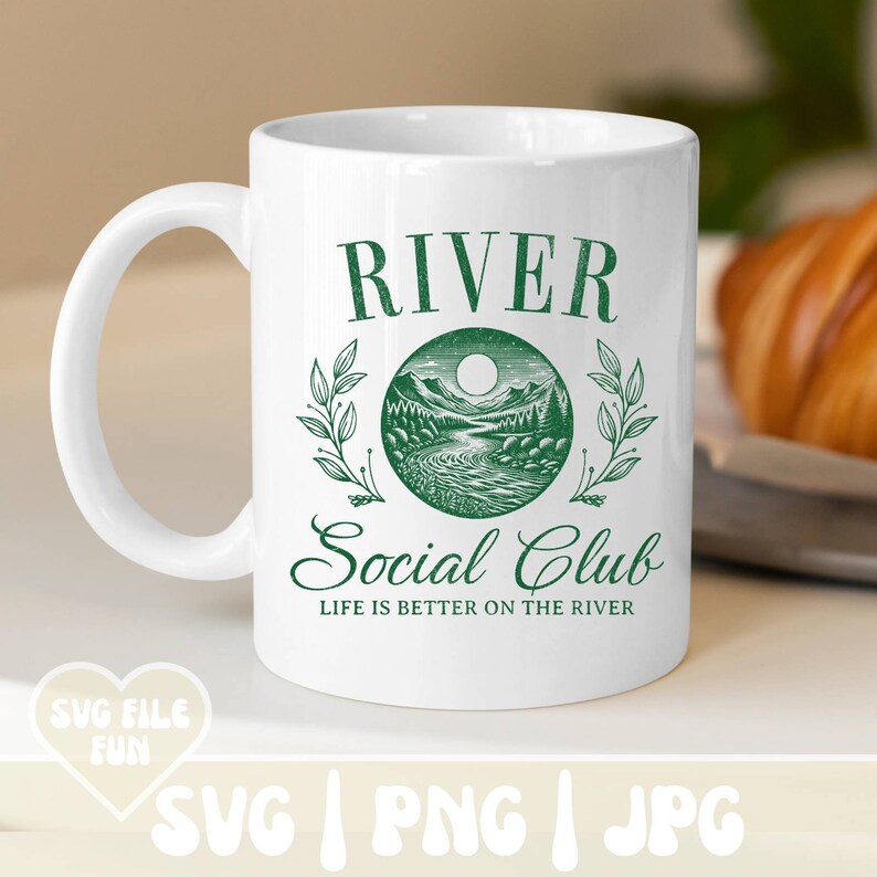 May include: White ceramic mug with a green graphic that reads "RIVER Social Club" with a river scene illustration. The text "LIFE IS BETTER ON THE RIVER" is also included. A croissant is in the background.