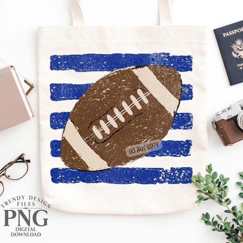 K&ouml;nnte beinhalten: Eine cremefarbene Canvas-Tragetasche mit einer braunen American-Football-Grafik, die &uuml;ber horizontalen, abgenutzten blauen Streifen zentriert ist. Der Football hat wei&szlig;e Schn&uuml;rsenkel und helle Endstreifen. Der Text DO NOT COPY ist auf dem Ball zu sehen.