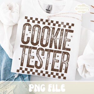 May include: White sweatshirt with the text "COOKIE TESTER" in brown, with a checkered pattern above and below the text. The words are in a bold, distressed font, with small star accents. The phrase "PNG FILE" is at the bottom.