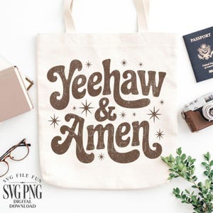 Puede incluir: Un bolso tote de color crema con las palabras "Yeehaw & Amen" en marr&oacute;n, con letras de estilo retro. El dise&ntilde;o incluye estrellas. La bolsa est&aacute; rodeada por un pasaporte, una c&aacute;mara vintage y otros accesorios.