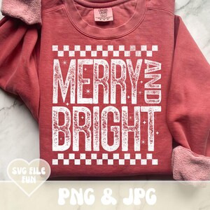 May include: A red-orange sweatshirt with the words "MERRY AND BRIGHT" in large white letters. The text is framed with a white checkered pattern. The sleeves are cuffed with a pink fabric.