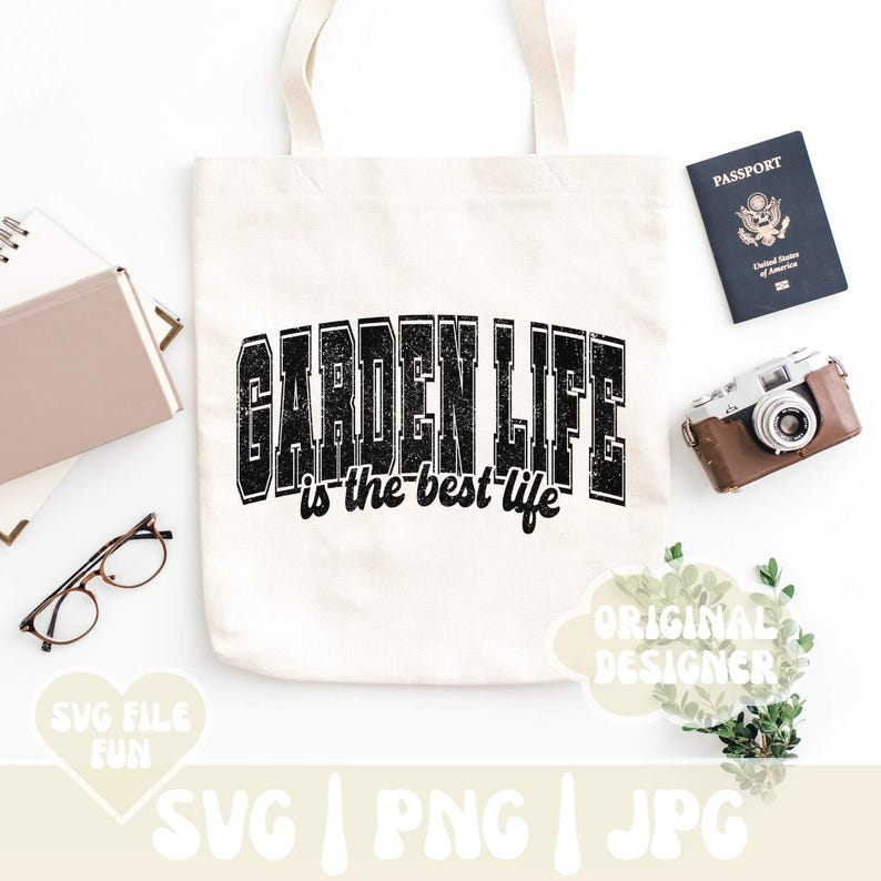 May include: A white tote bag with black text that reads "Garden Life is the best life". The bag is on a white surface with a passport, camera, and plants in the background.