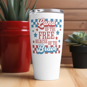 May include: A white tumbler with a lid, featuring the text "Land of the Free Because of the Brave" in red and blue. The design includes stars and a checkered pattern, evoking patriotic themes. The tumbler is displayed on a wooden surface.