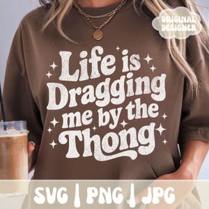 May include: A brown sweatshirt with the phrase "Life is Dragging me by the Thong" in white, stylized text. The design includes star accents. The bottom of the image shows "SVG | PNG | JPG".