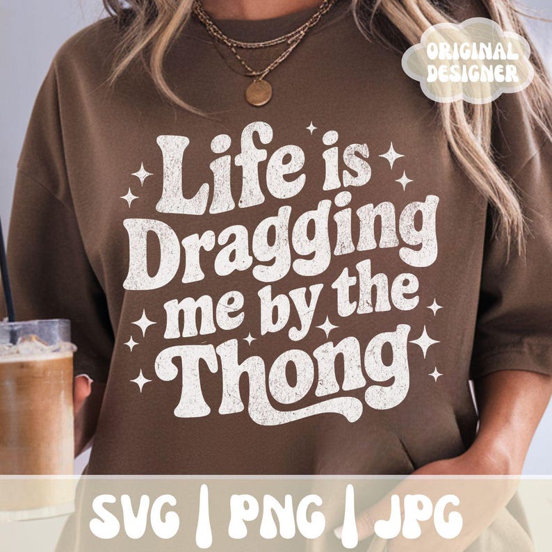 May include: A brown sweatshirt with the phrase "Life is Dragging me by the Thong" in white, stylized text. The design includes star accents. The bottom of the image shows "SVG | PNG | JPG".