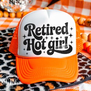 May include: A bright orange trucker hat with a white front panel featuring the black text Retired Hot girl! in a stylised font, accented by small black star shapes. The hat has a structured foam front and a mesh back with an adjustable snap closure.