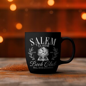 K&ouml;nnte beinhalten: Schwarze Keramiktasse mit einem wei&szlig;en Design, auf dem "Salem Witch Book Club Est. 1692 Bell, Book & Candle" steht. Das Design zeigt ein Buch mit einem Halbmond und Sternen dar&uuml;ber.