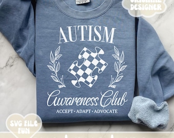 Op de afbeelding: Een blauw sweatshirt met witte tekst die "Autism Awareness Club" luidt met een wit puzzelstukje en een krans van bladeren. De tekst zegt ook "Accepteren, Aanpassen, Pleiten".