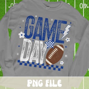 Puede incluir: Una camiseta gris de manga larga con la frase "GAME DAY" en azul y blanco, con un gr&aacute;fico de f&uacute;tbol americano. El dise&ntilde;o incluye una estrella a cuadros azul y blanco, un rayo y detalles florales. La camiseta es un archivo PNG.