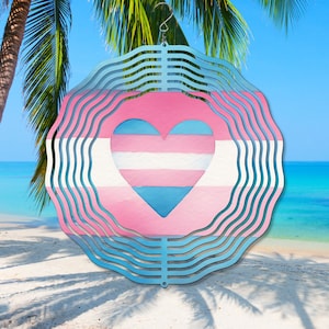 May include: A metal wind spinner with a layered design featuring a heart in the center. The heart is painted in the colors of the transgender pride flag: light blue, pink, and white. The wind spinner is suspended from a silver hook.