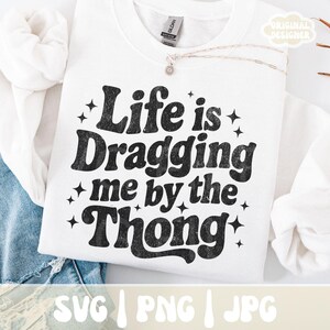 May include: White sweatshirt with the phrase "Life is Dragging me by the Thong" in black, retro-style lettering. The design includes star accents. The sweatshirt is displayed on a white surface with a denim garment visible.