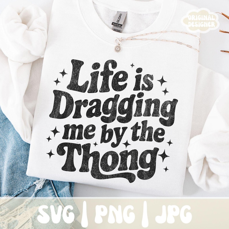 May include: White sweatshirt with the phrase "Life is Dragging me by the Thong" in black, retro-style lettering. The design includes star accents. The sweatshirt is displayed on a white surface with a denim garment visible.