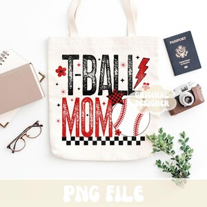 May include: A white tote bag with the text "T-BALL MOM" in black and red, featuring a baseball graphic. The bag is surrounded by a passport, a camera, and other accessories.