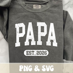 以下が含まれることがあります： グレーのComfort Colorsスウェットシャツ。大きな白い文字で「PAPA」と書かれています。下には、白い楕円の中に「EST. 2026」という文字があります。画像の下部には「PNG & SVG」という文字が含まれています。