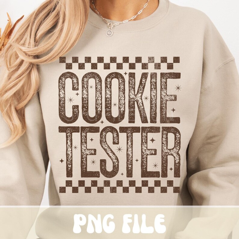 May include: Beige sweatshirt with the text "COOKIE TESTER" in a brown, distressed font. The text is framed by a brown and white checkered pattern. The words are decorated with small starbursts.