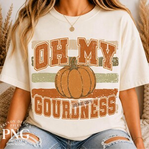 May include: A cream-colored short-sleeve t-shirt featuring a central orange pumpkin graphic. The text reads OH MY GOURDNESS in bold, distressed orange block letters. The design is layered over horizontal stripes in muted orange and sage green tones.