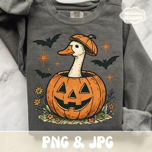 May include: Dark gray sweatshirt with a Halloween-themed graphic. The design features a white duck wearing a pumpkin hat, emerging from an orange jack-o'-lantern. Bats, stars, and flowers surround the pumpkin. The text "PNG & JPG" is at the bottom.