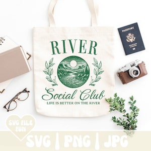 May include: Cream-colored tote bag with a green graphic that reads "RIVER Social Club" and "Life is better on the river." The design features a river scene. A passport, camera, and greenery are also present.