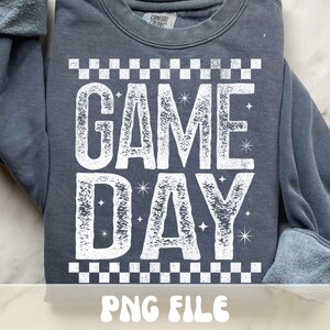 K&ouml;nnte beinhalten: Ein dunkelgraues Sweatshirt mit dem Schriftzug "GAME DAY" in gro&szlig;en, verwaschenen wei&szlig;en Buchstaben. Das Design umfasst ein Schachbrettmuster &uuml;ber und unter dem Text, mit Sternen. Der untere Teil des Bildes zeigt "PNG FILE".