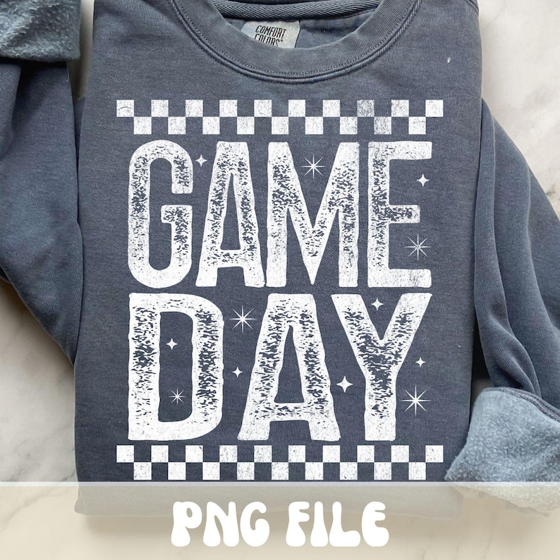 K&ouml;nnte beinhalten: Ein dunkelgraues Sweatshirt mit dem Schriftzug "GAME DAY" in gro&szlig;en, verwaschenen wei&szlig;en Buchstaben. Das Design umfasst ein Schachbrettmuster &uuml;ber und unter dem Text, mit Sternen. Der untere Teil des Bildes zeigt "PNG FILE".