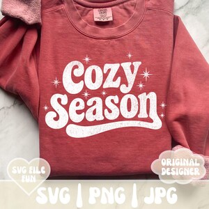 Puede incluir: Una sudadera de color rojo anaranjado con la frase "Cozy Season" en blanco, con letras de estilo retro. El dise&ntilde;o incluye detalles de estrellas. La sudadera tiene cuello redondo y mangas largas. La imagen tambi&eacute;n incluye el texto "SVG FILE FUN" y "ORIGINAL DESIGNER".